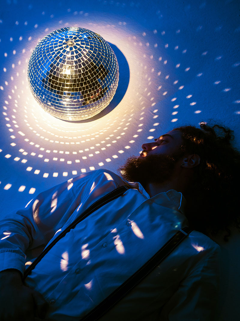 photo of man lying down and looking at reflective spinning ball with spots of light reflected onto him and the floor