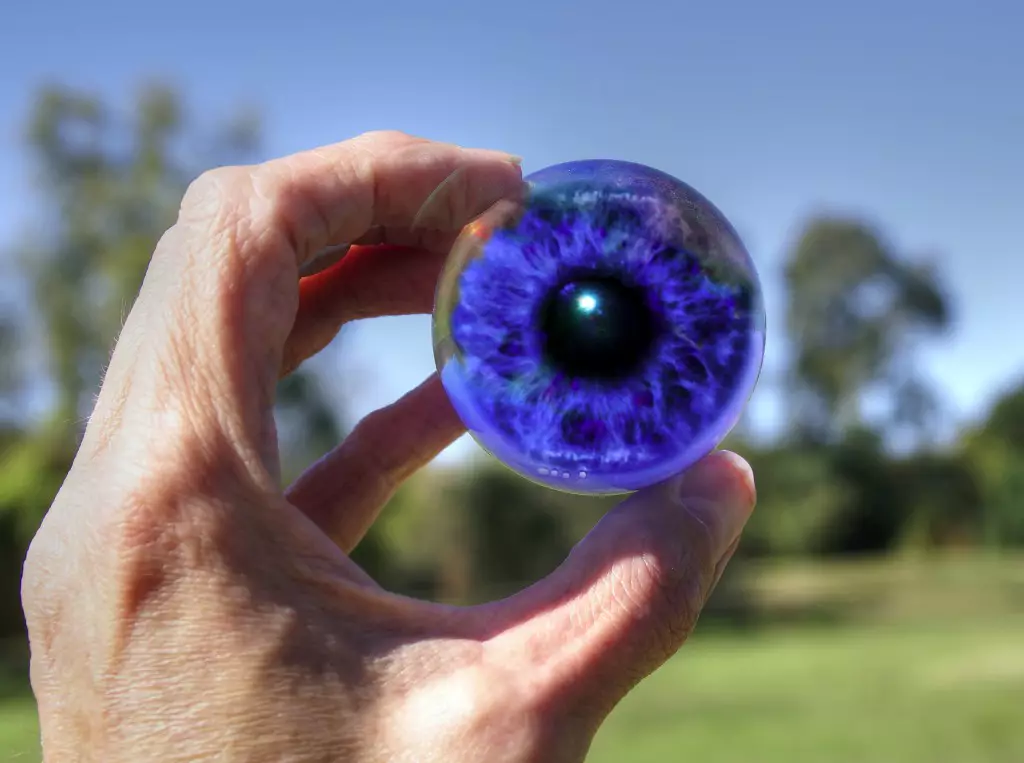 eye of blue photoshopped into glass sphere being held in fingertips in front of park scene