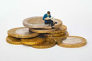 small figure of man sits on pile of big coins reading a thick newspaper